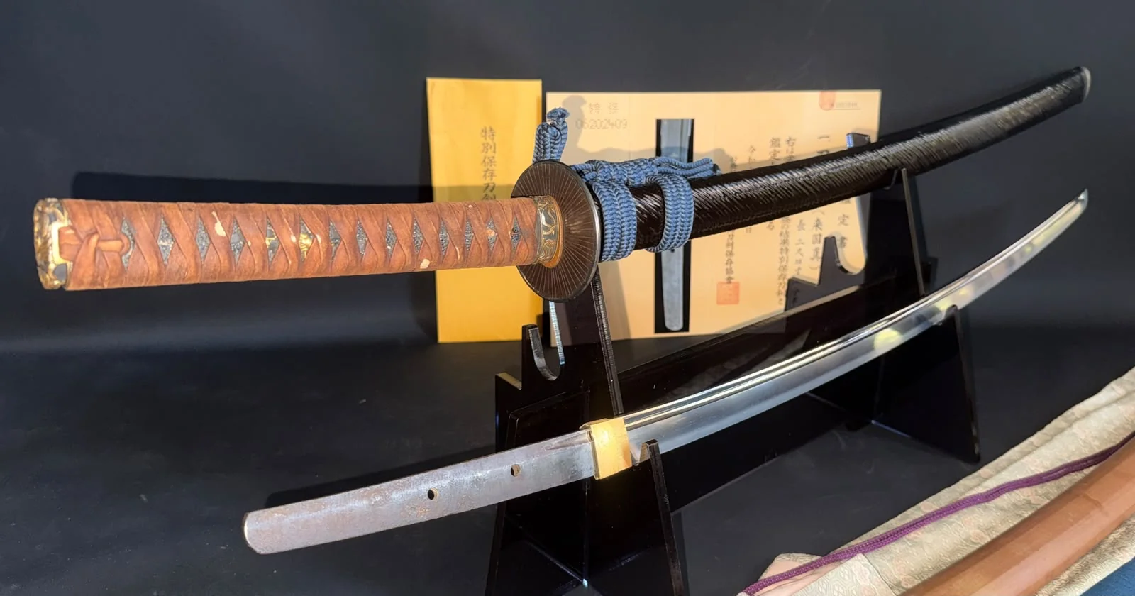 Side view of the Rai Kunizane katana koshirae showing the black lacquer saya, brown ito wrap, blue sageo and graceful late Kamakura curvature of the polished blade